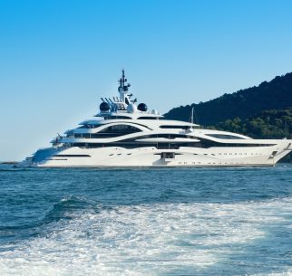 Huge luxury motor yacht cruising offshore in a calm ocean passing a forested coastline viewed over the wake of a passing ship